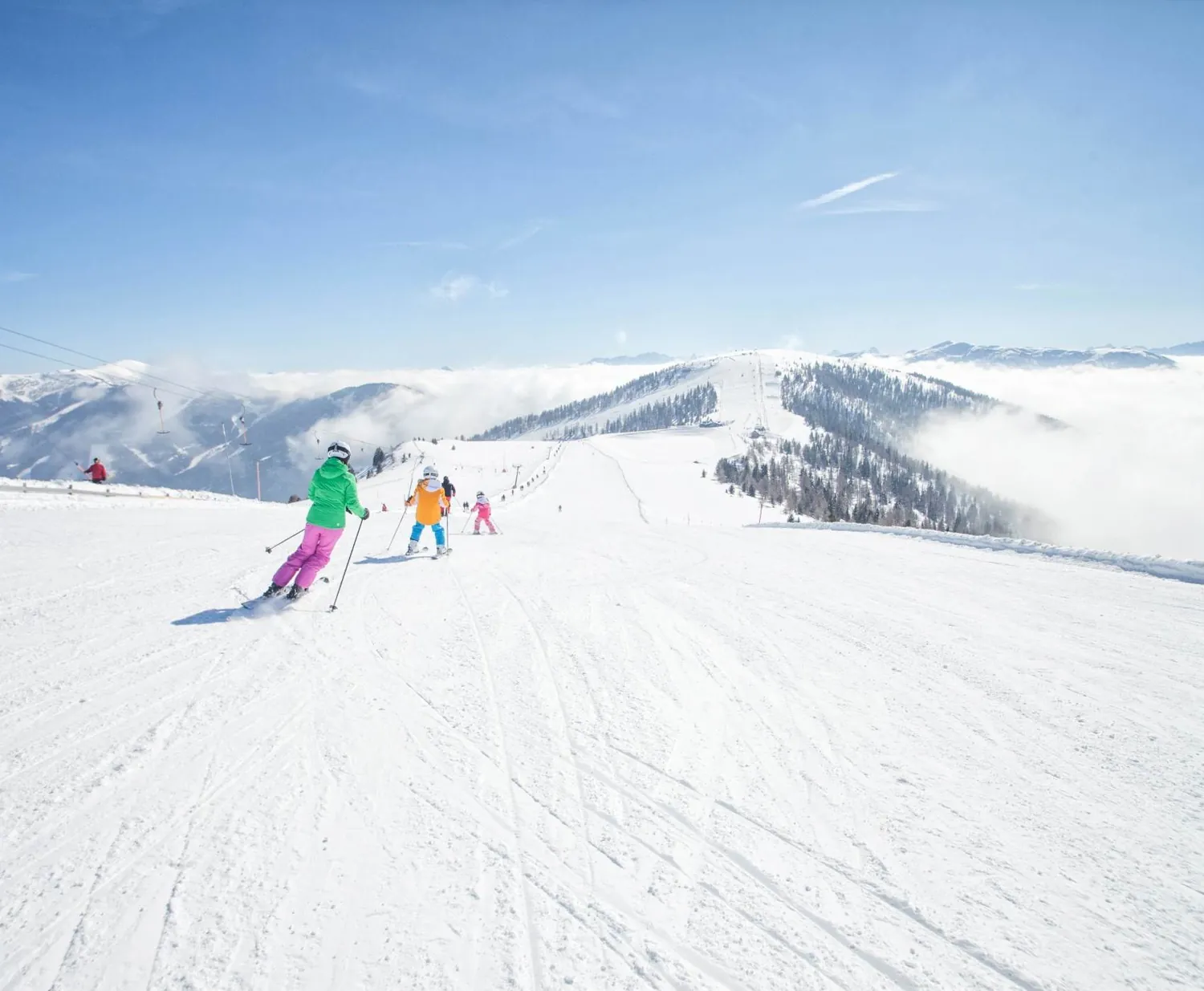 Skifahrer in bunter Kleidung auf breiter Piste über dem Wolkenmeer – Skifahren mit Familie und Freunden
