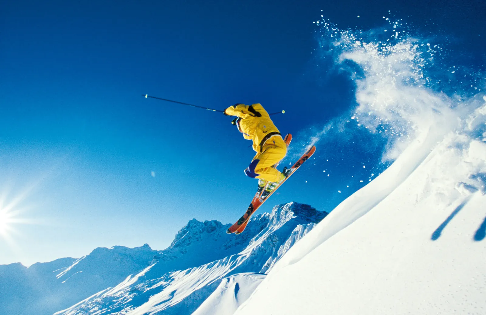 Sprung im Tiefschnee Skifahrer im gelben Anzug im Sprung über eine Schneekante vor Gipfeln und tiefblauem Himmel – Freeride und Skispaß