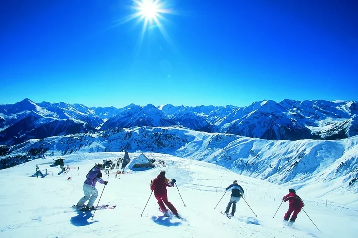 Vier Skifahrer auf der Abfahrt vor einem weiten Bergpanorama unter blauem Himmel – Skiurlaub mit Freunden