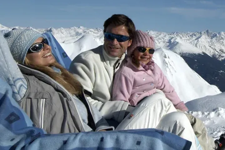 Familie mit Kind macht Pause in der Sonne vor einem weiten Bergpanorama – Familienskiurlaub