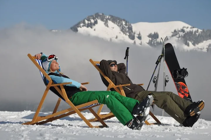 Paar entspannt im Liegestuhl neben Skiern und Snowboard über den Wolken – Pause im Skiurlaub