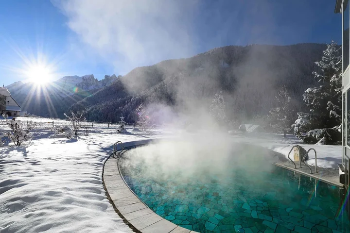 Beheizter Außenpool im Schnee mit aufsteigendem Dampf und Felsgipfeln im Gegenlicht – Auszeit nach dem Skifahren