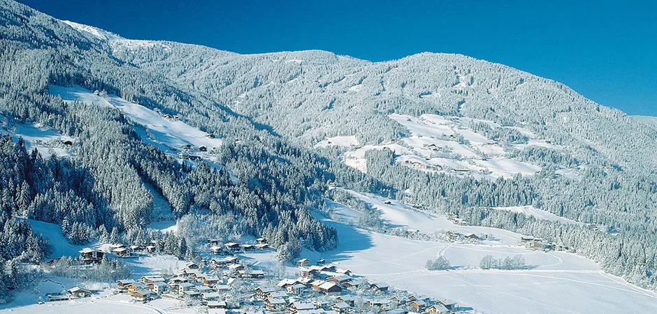 Verschneiter Bergort zwischen Tannenwäldern bei klarem Himmel – Winterurlaub in den Bergen
